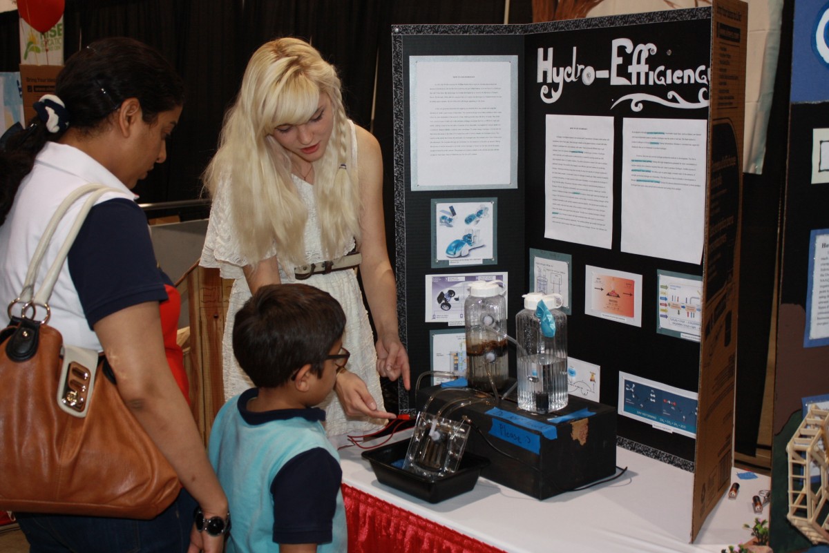 Una chica explica un invento sobre el agua a una madre y su hijo en una feria de ciencias. 