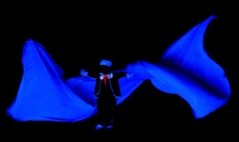 Sobre un escenario totalmente oscuro se aprecia el contorno de una figura delante de una tela azul.
