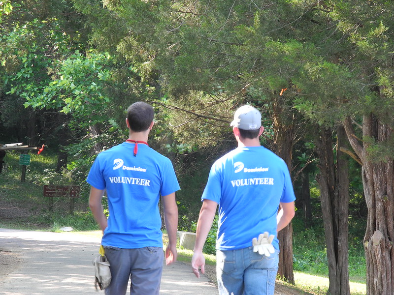 En la imagen puedes ver dos voluntarios con camiseta celeste caminando junto a un bosque