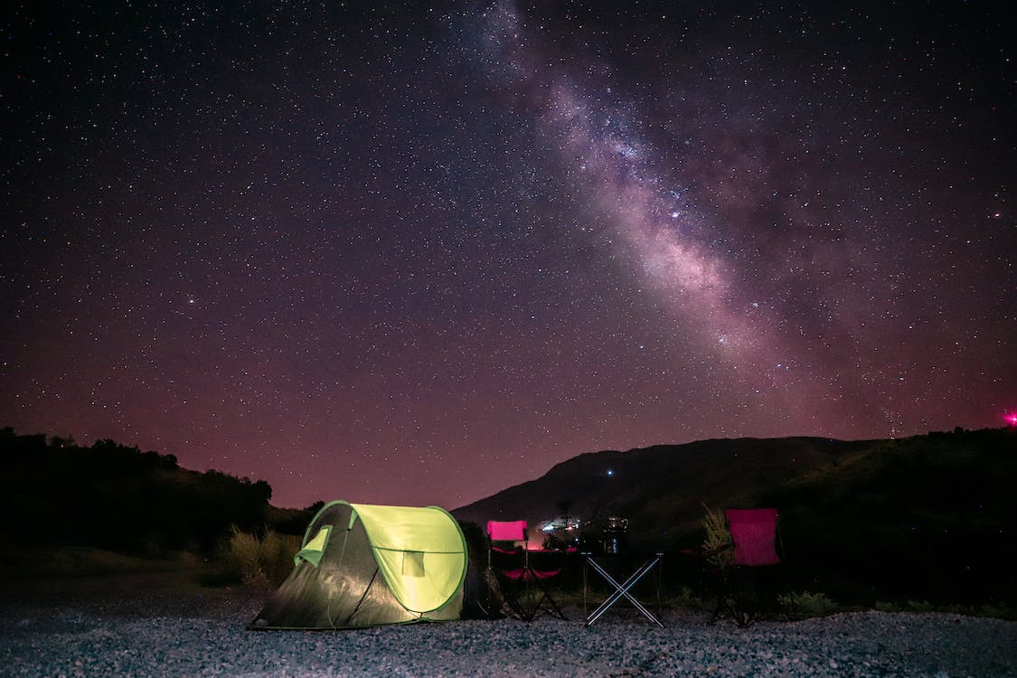 La imagen muestra una tienda de campaña verde y unas mesas y sillas bajo el cielo estrellado de noche en una llanura en el campo.