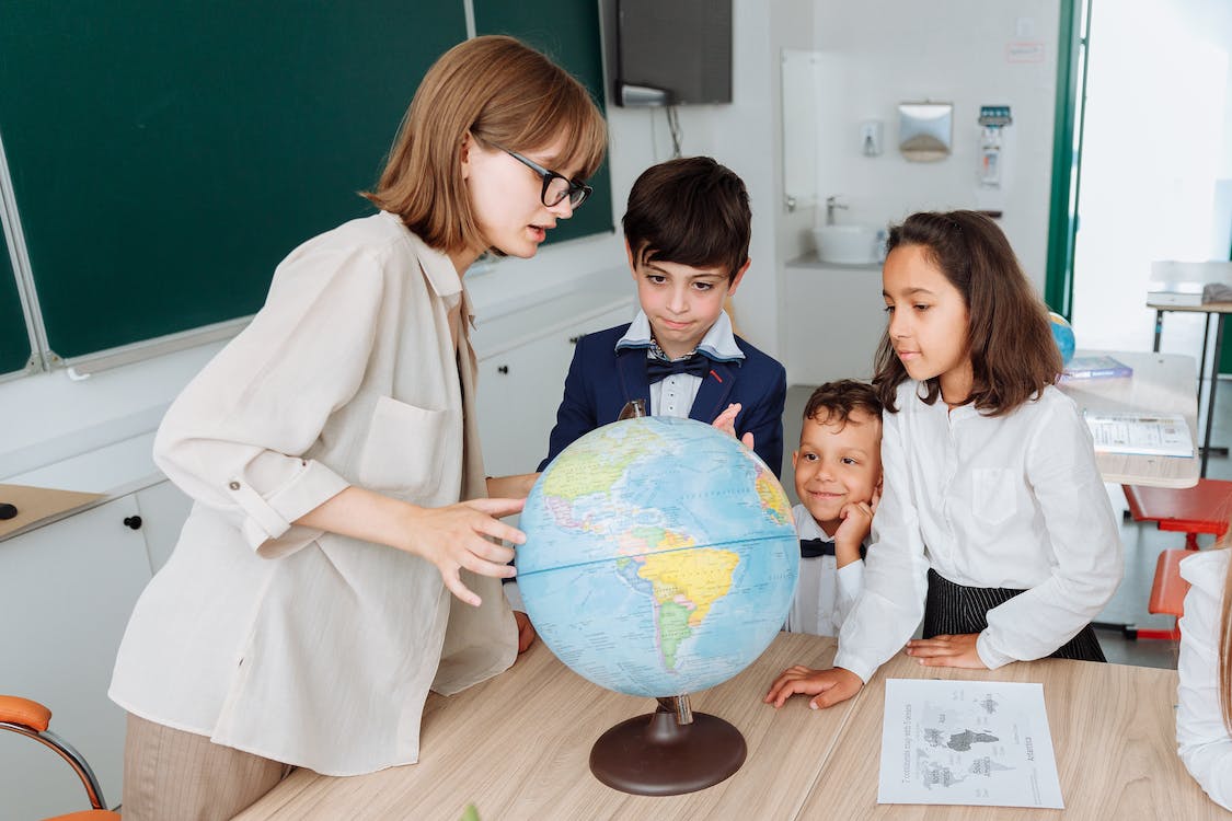 La imagen muestra una clase en un colegio y hay una profesora mirando a un globo terráqueo mientras habla y tres niños observando atentamente.