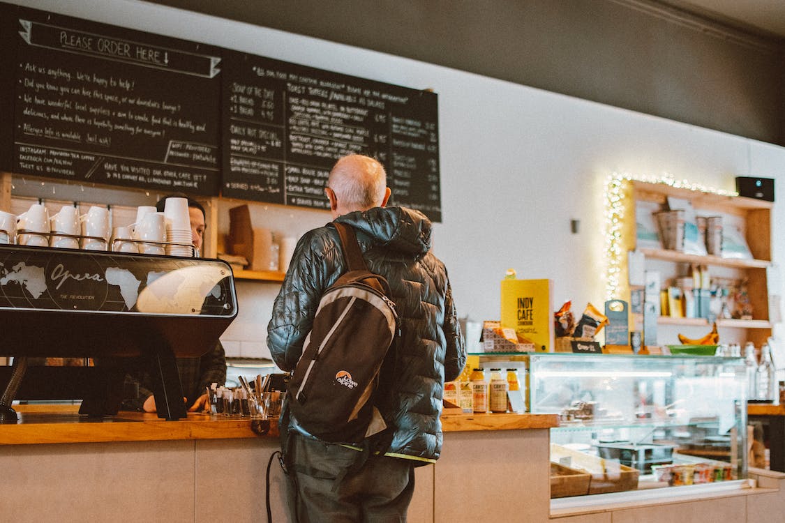 La imagen muestra un cliente de espalda en el mostrador de una cafetería mientras es atendido.