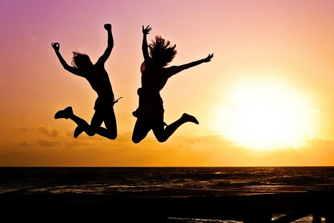 La imagen muestra a dos chicas saltando con los brazos levantados en una playa al atardecer. 