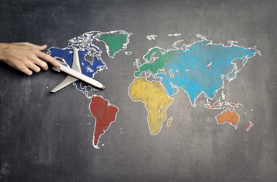 La imagen muestra un mapa del mundo dibujado en colores sobre una pizarra y sobre la que hay un avión sujetado por una mano. 