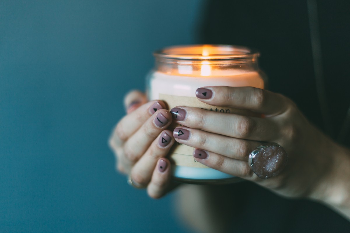 En la imagen puedes ver unas manos con las uñas pintadas sujetando una vela encendida en un tarro de cristal