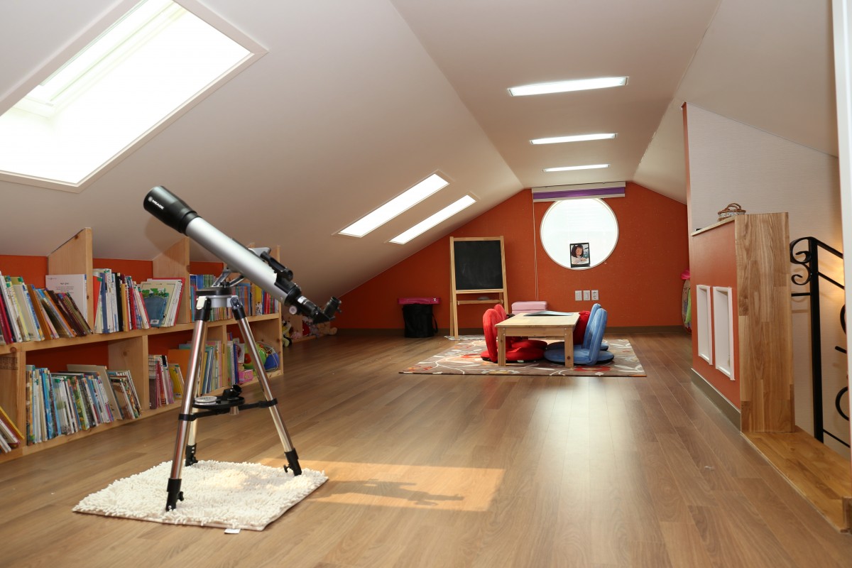 En la imagen aparece un ático con una librería, un telescopio y al fondo una mesa, y una pizarra pequeña, junto a un tragaluz