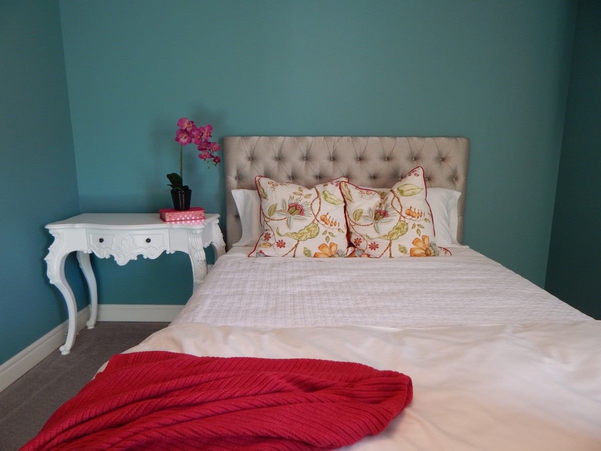 n la imagen se ve una cama con sábanas blancas y una manta roja encima, además de dos cojines estampados
