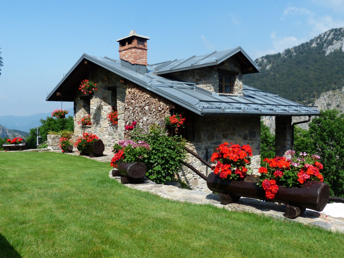 En la imagen puedes ver una casa aislada en el campo que tiene varios maceteros con flores y el suelo de hierva a su entrada. Al fondo se ven unas montañas