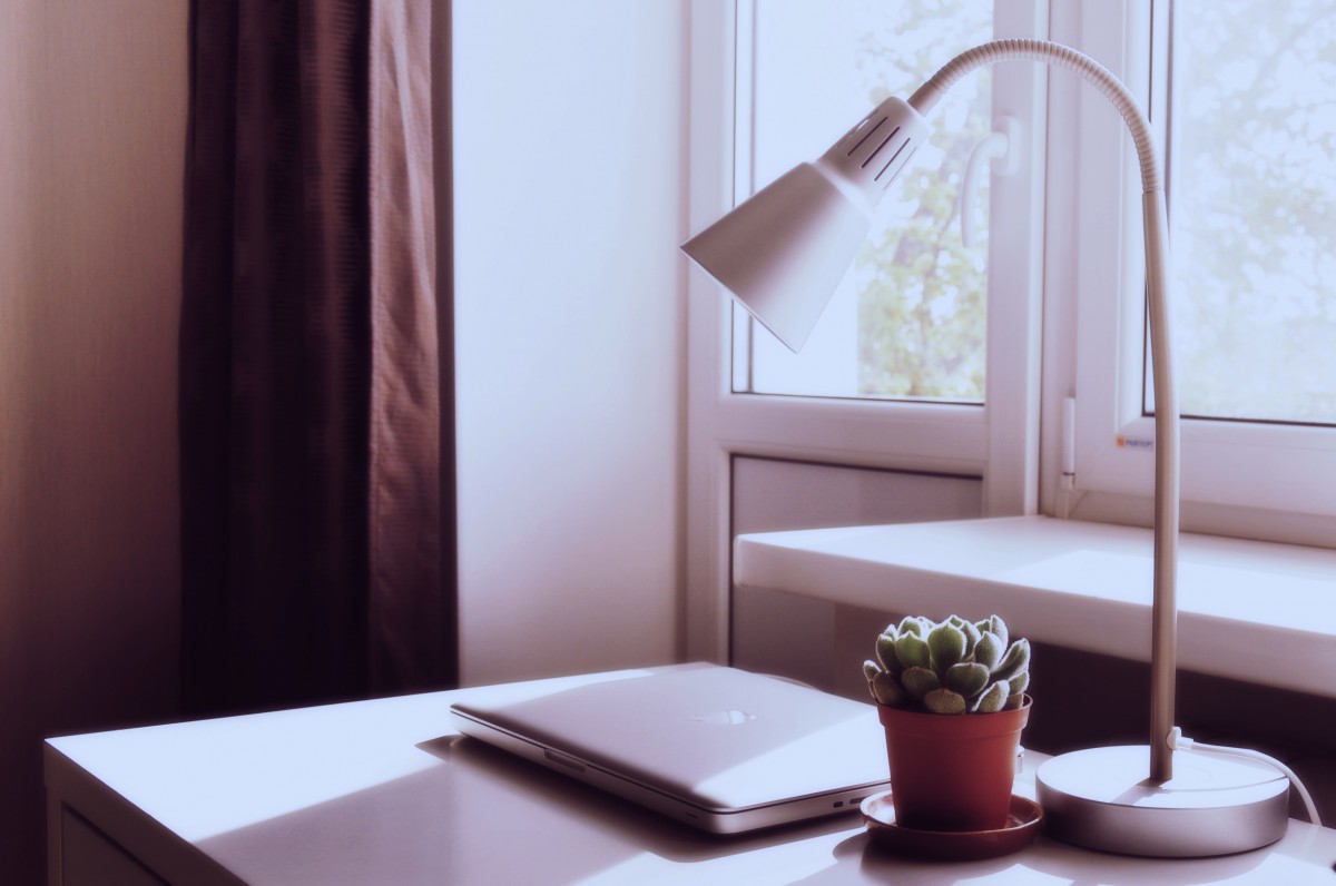En la imagen puedes ver una mesa de escritorio con un ordenador, una planta pequeña y una lámpara tipo flexo