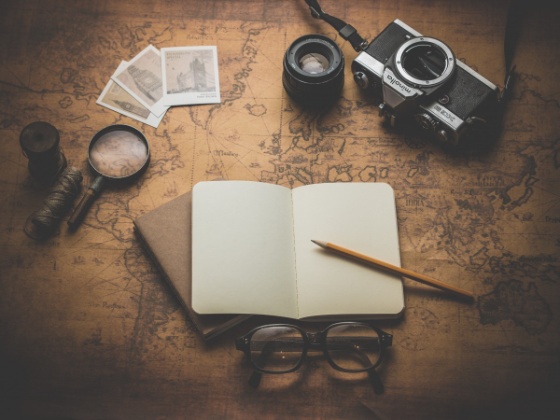 La imagen muestra una libreta, lápiz, cámara de fotos, gafas y demás utensilios para planificar un viaje