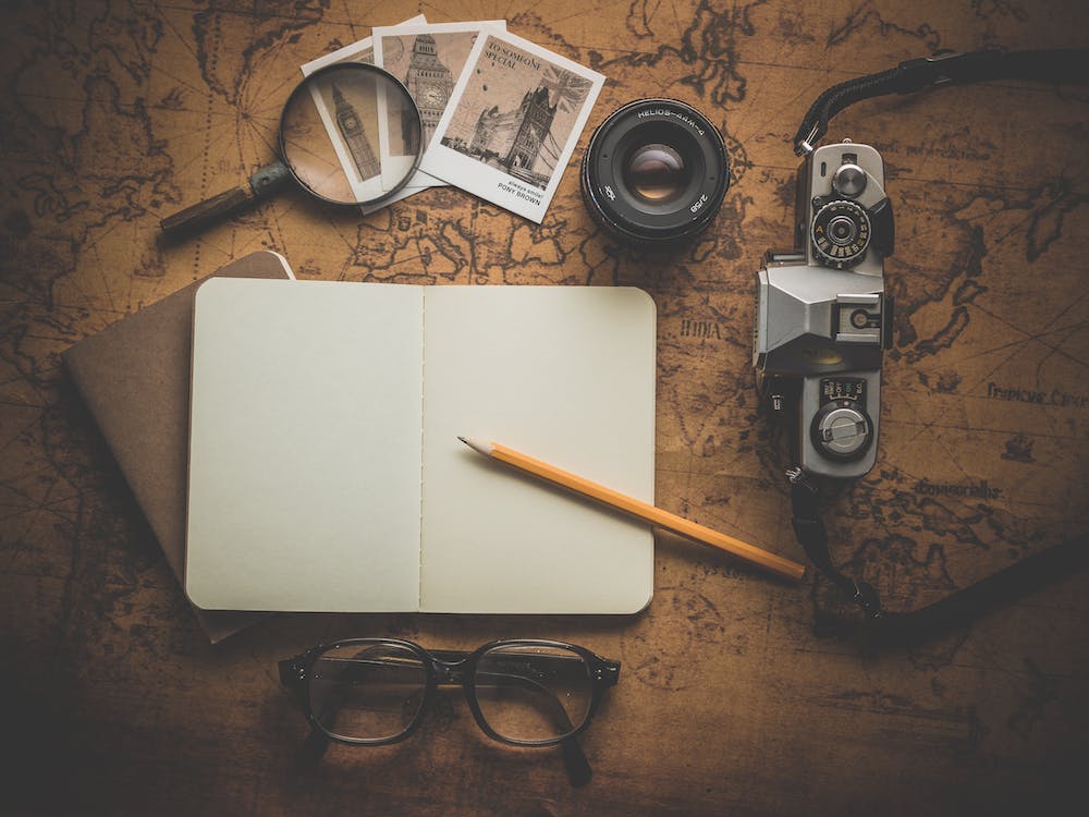 La imagen muestra un libro en blanco abierto, un lápiz, una cámara de fotos, una lupa y unas fotografías sobre una mesa de escritorio marrón. 