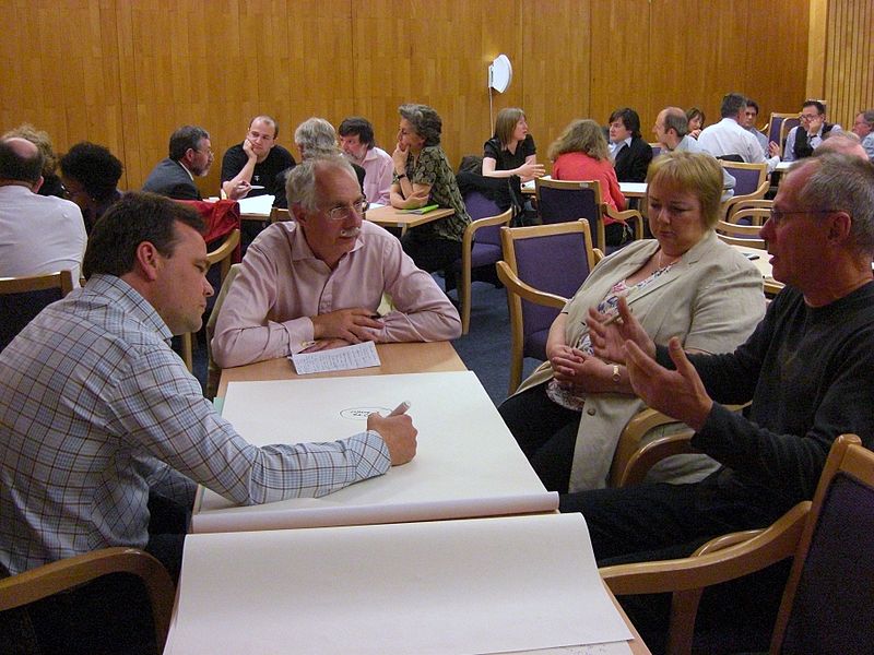 La imagen muestra en primer plano a un grupo de cuatro personas sentados en una mesa mientras charlan y detrás de ellos hay más grupos de personas que también se encuentran charlando.