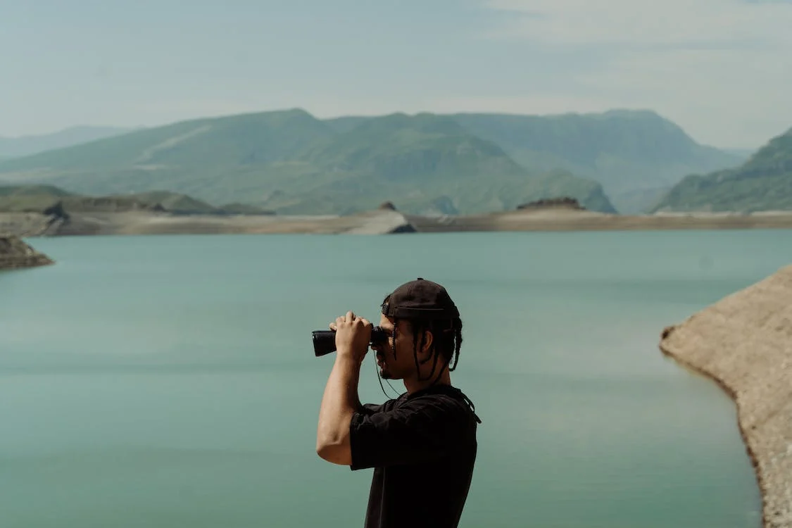 La imagen muestra a un hombre con camiseta y gorro negra mirando a través de unos prismáticos el paisaje de un lago turquesa con montañas a lo lejos. 