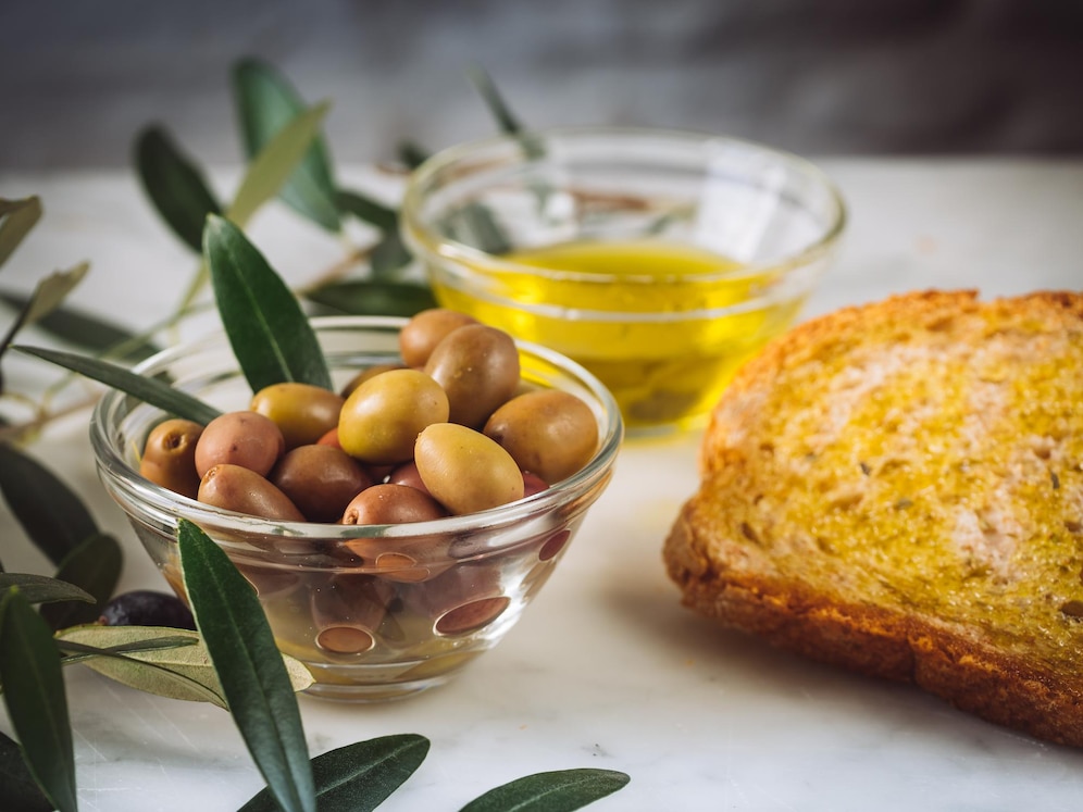 La imagen muestra una tostada de pan con aceite de oliva, un cuenco con aceitunas y ramas de olivo y en el fondo un cuenco con aceite de oliva.  