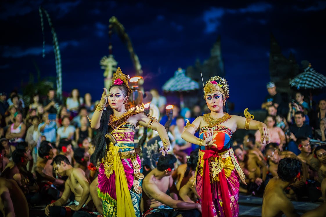 La imagen muestra en primer plano a dos mujeres bailarinas vestidas con ropa típica asiática de colores llamativos por la noche.