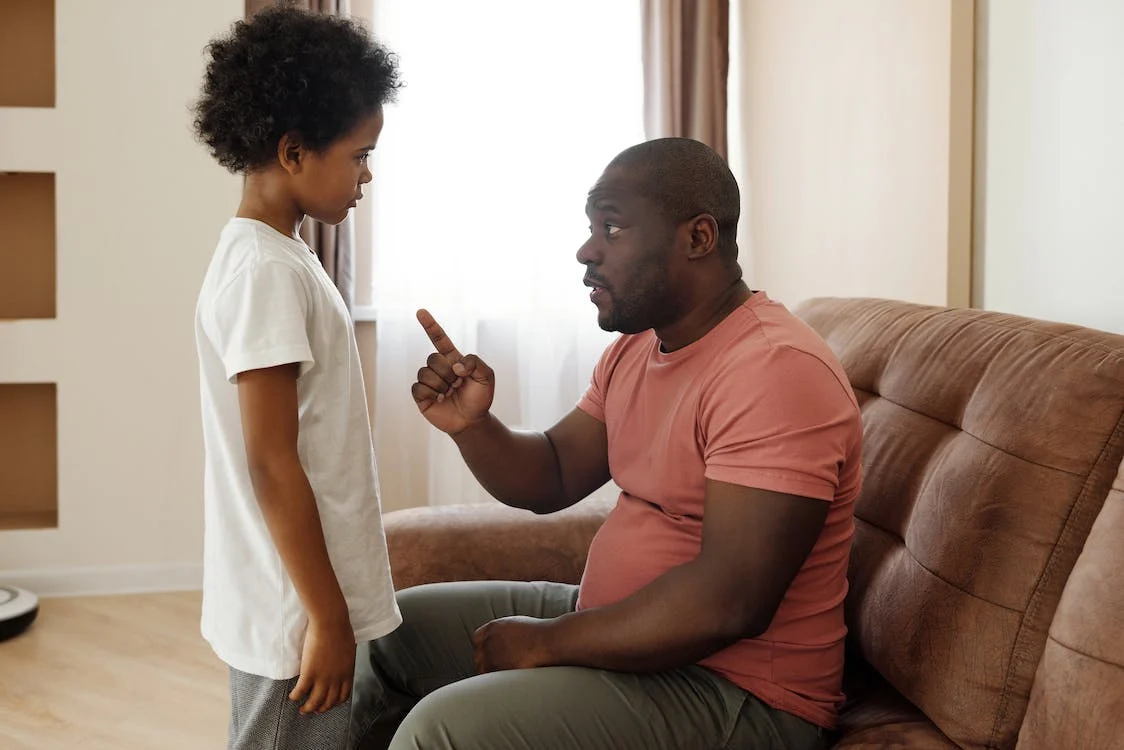 La imagen muestra a un padre sentado en el sofá mientras riñe con el dedo índice dirigiéndose a su hijo que lo está escuchando atentamente por haberse portado mal.