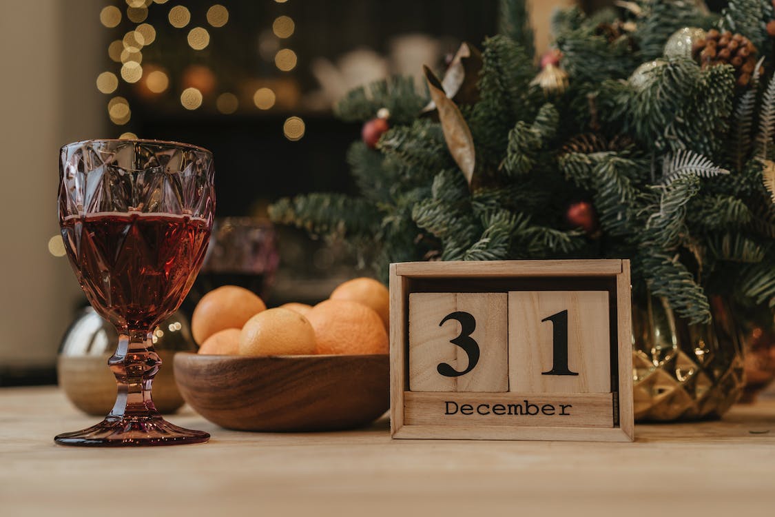 La imagen muestra un calendario de madera que muestra el número treinta y uno de diciembre sobre una mesa, a la izquierda una copa de vino tinto y al fondo adornos de navidad. 