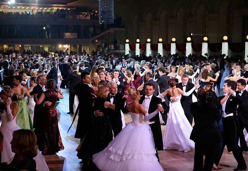 La imagen muestra un salón de baile en el que se encuentran muchas parejas, vestidas formalmente para la ocasión, bailando.