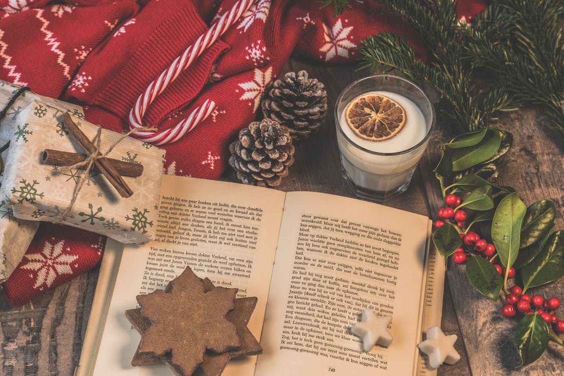 La imagen muestra una mesa decorada con adornos navideños, hojas de árboles, caramelos en forma de bastón rojos y blancos, un pequeño regalo envuelto y un mantel rojo. En en centro de la imagen se muestra un libro abierto y arriba de este un vaso de leche con una rodaja de naranja dentro. 