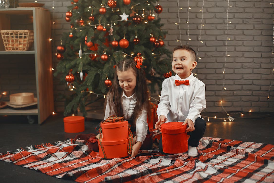 La imagen muestra a un niño y una niña en el salón decorado con motivos navideños. A la izquierda de la imagen aparece un árbol de navidad decorado con luces y los niños están abriendo unos regalos de color rojo.