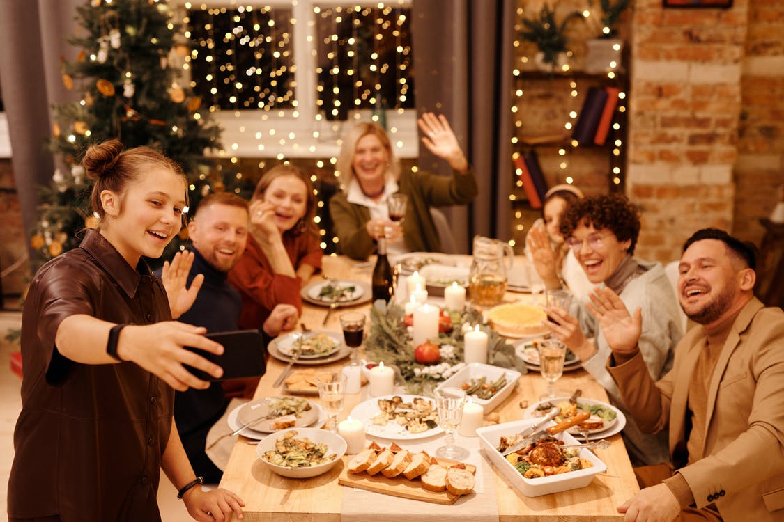La imagen muestra a una familia que están alrededor de una mesa dispuestos a cenar en una habitación decorada con adornos de navidad y mirando a un móvil que está sujetando una mujer a la izquierda para hacerse una foto. 