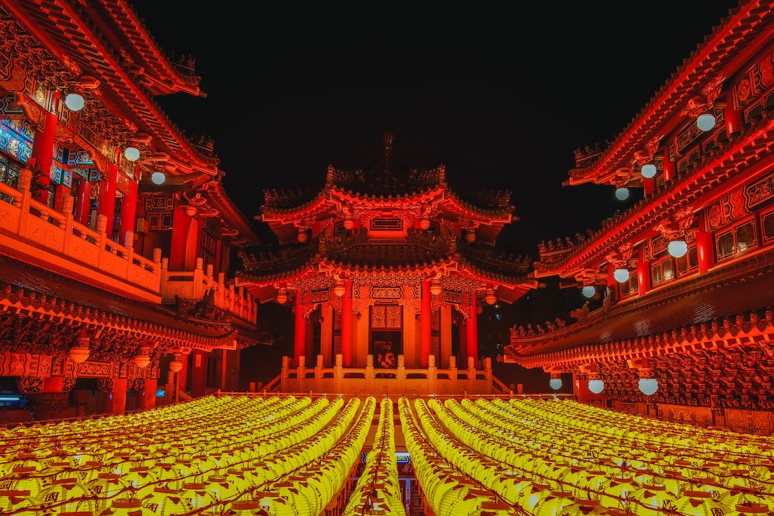 La imagen muestra un edificio grande típico chino con tejados puntiagudos de color rojo de noche. A ambos lados aparecen dos edificios de color rojo decorados con adornos chinos. En la parte inferior aparece varias líneas de farolillos chinos encendidos de color amarillo y brillantes. 