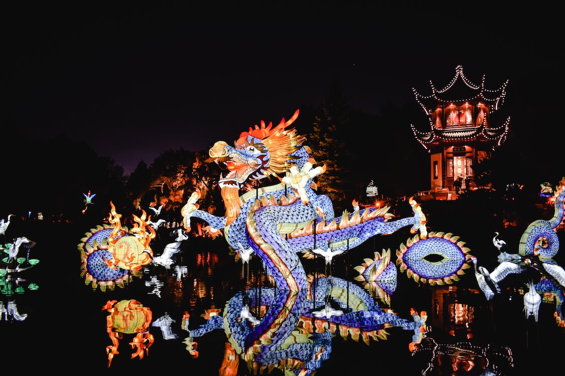 La imagen muestra a un dragón típico chino hecho de telas brillantes de color azul y bailando por las calles sobre un cielo negro con un edificio chino con techos puntiagudos al fondo de la imagen. 