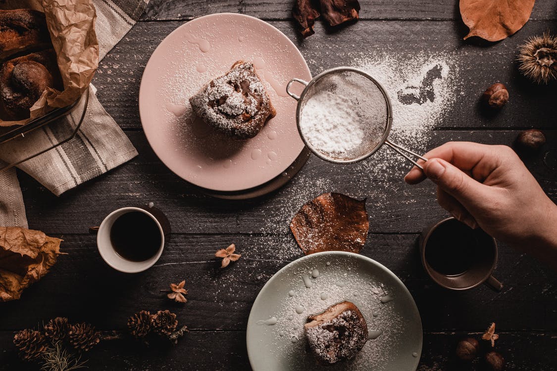 La imagen muestra una mesa marrón vista desde arriba decoara con piñas, tazas de cafés y dos platos con dos trozos de bizcochos. A la derecha una mano sujeta un colador espolvoreando sobre los postres azúcar en polvo.