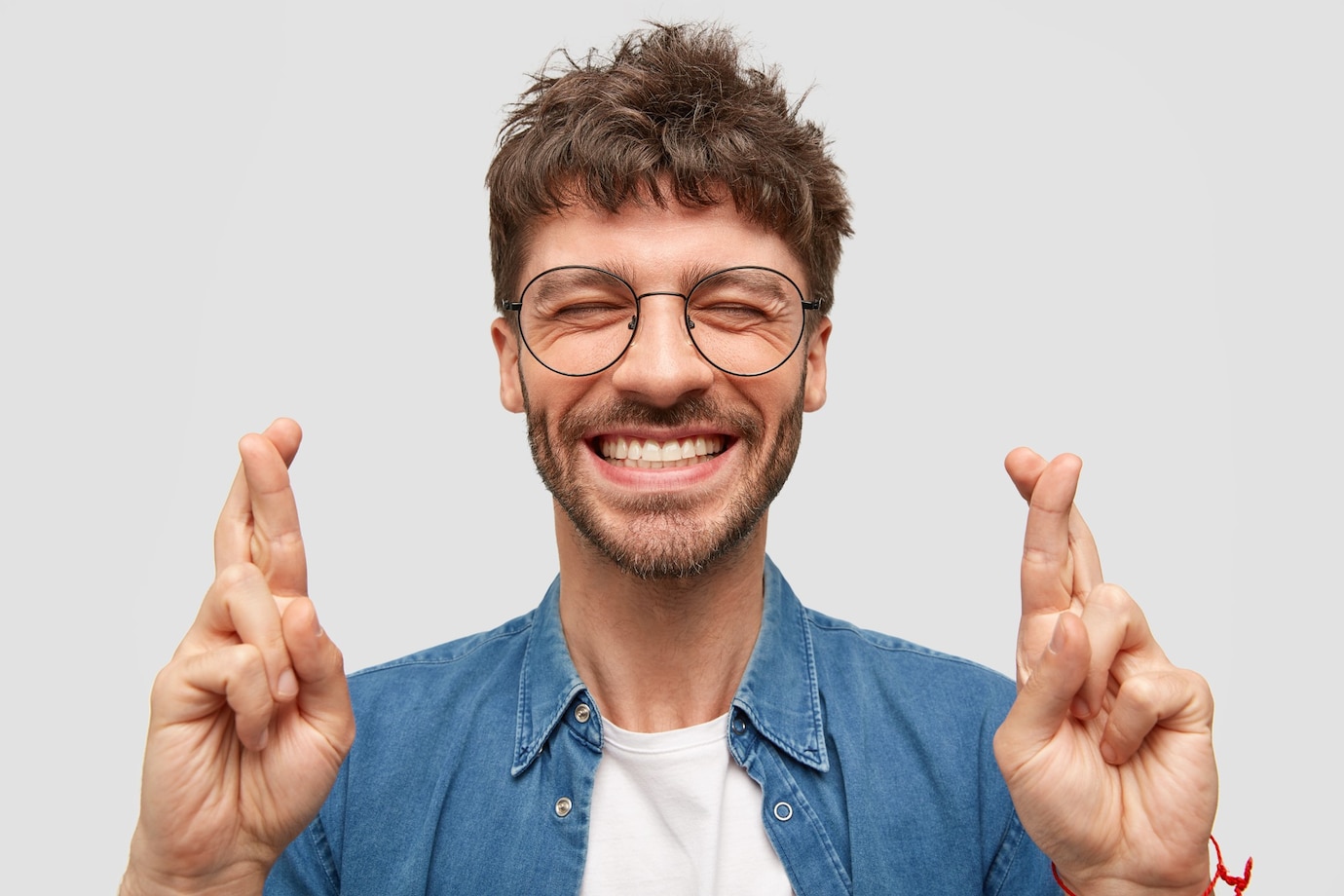 La imagen muestra a un hombre con barba y gafas con los ojos cerrados, sonriendo y con los dos dedos de las dos manos cruzados a modo de buena suerte.