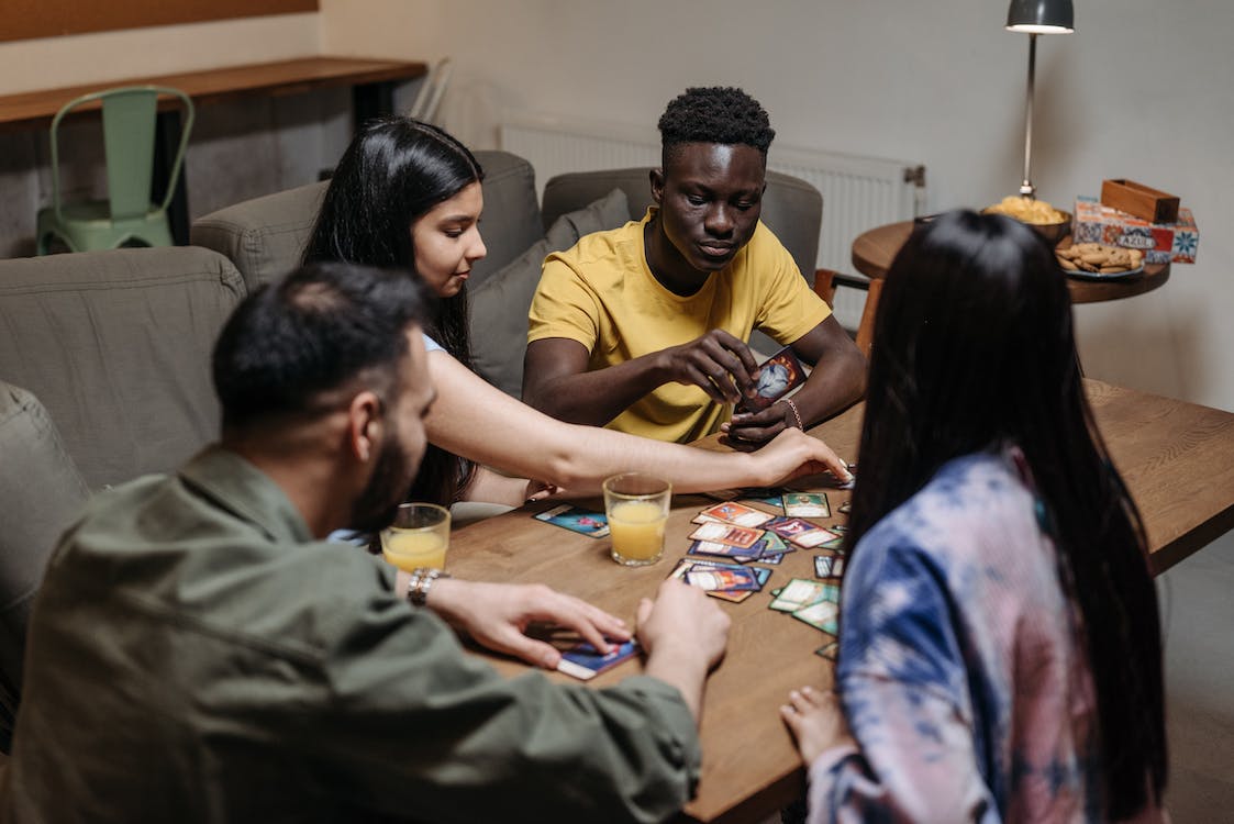 La imagen muestra cuatro personas alrededor de una mesa preparando un juego de mesa de cartas y encima de la mesa hay dos zumos de naranja.  