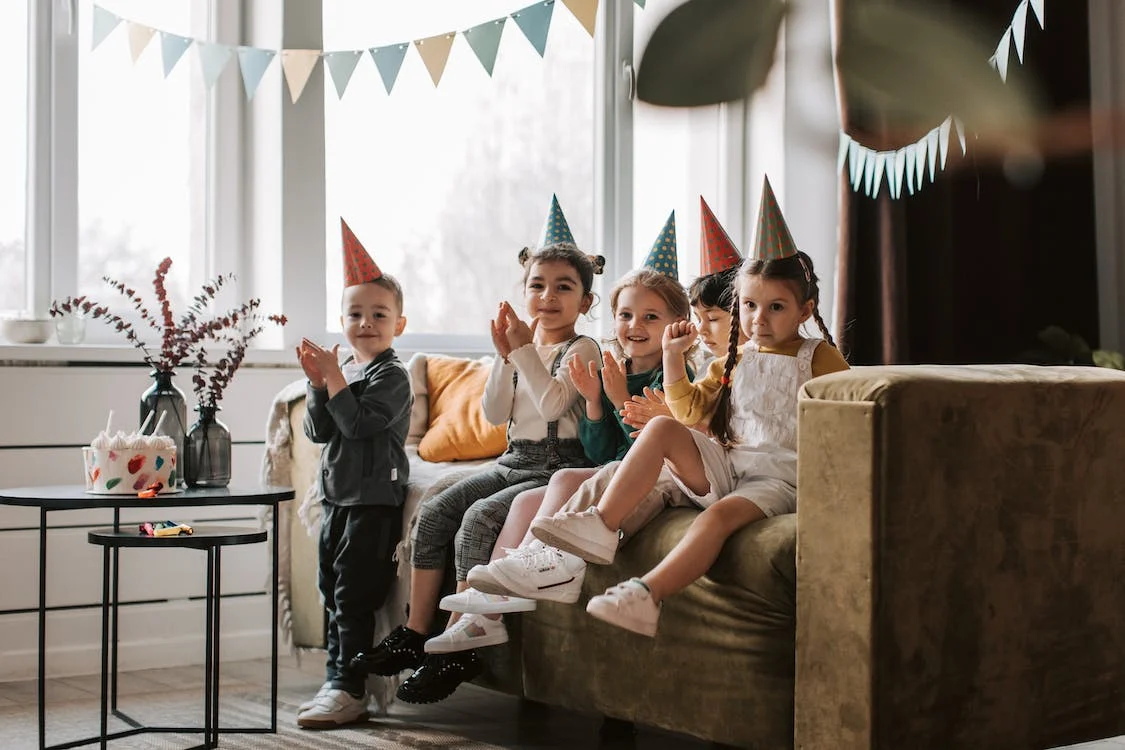 La imagen muestra a cinco niños en un salón portándose bien y sonriendo en una fiesta de cumpleaños. Los niños llevan puesto gorros de papel de cumpleaños y el salón está decorado con banderines de colores. 