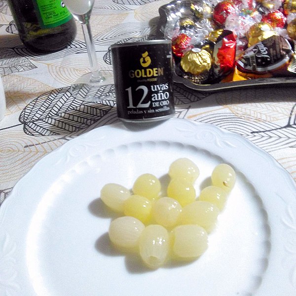  La imagen muestra una mesa con un plato blanco con doce uvas y una lata negra que se llama 12 uvas año de oro. A la derecha hay una bandeja con bombones y mantecados. 