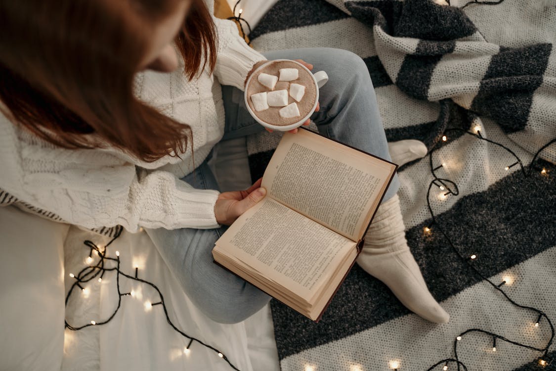 La imagen muestra desde arriba a una mujer sentada con las piernas cruzadas en la cama leyendo con un libro abierto en la mano derecha y una taza de chocolate caliente con nubes en la mano izquierda. La cama está decorada con luces de navidad y una manta negra y blanca de rayas. 
