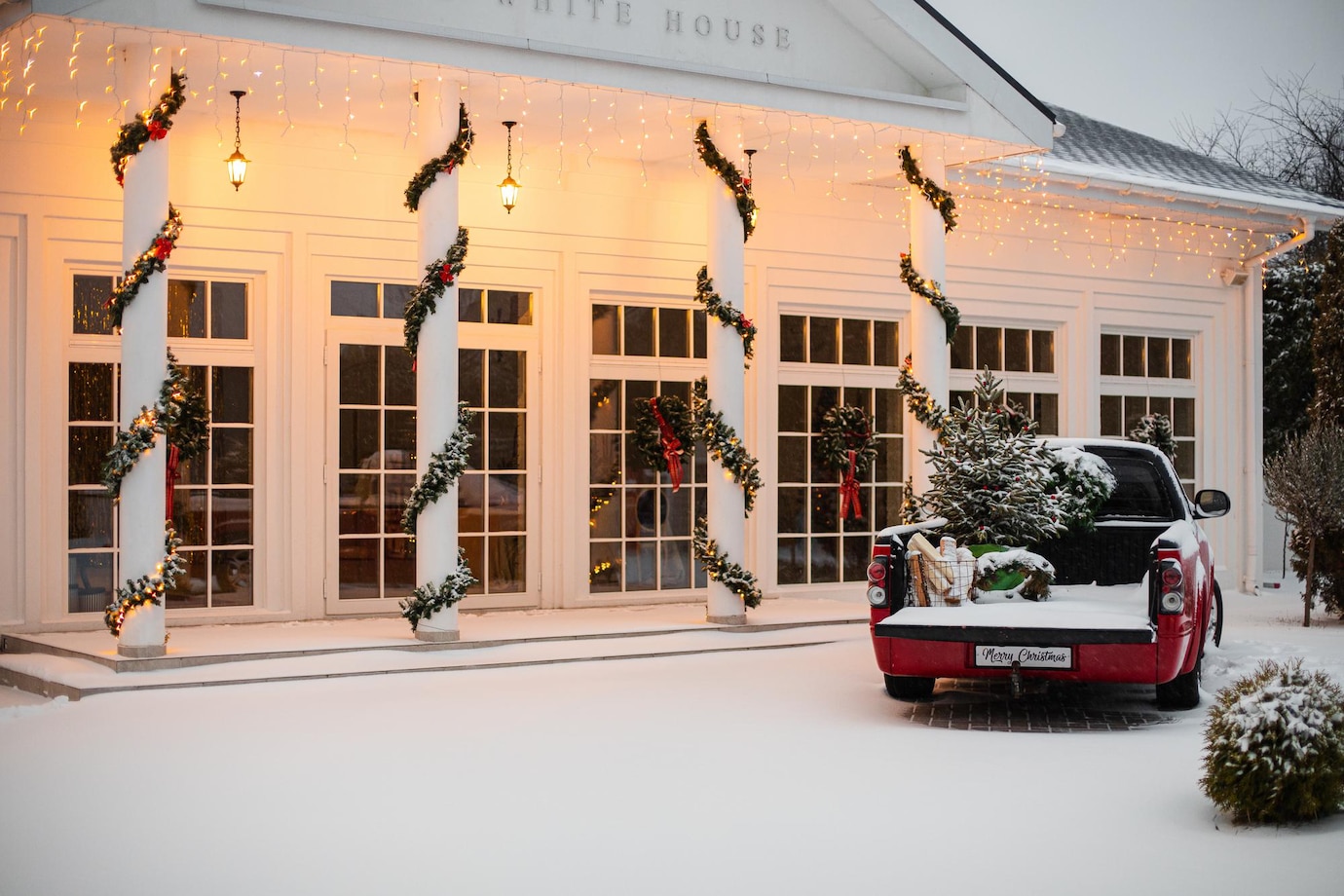 La imagen muestra una casa blanca con cuatro columnas decoradas con flores navideña. A la izquierda hay una caminoneta roja con un árbol de navidad sobre la calle nevada. 