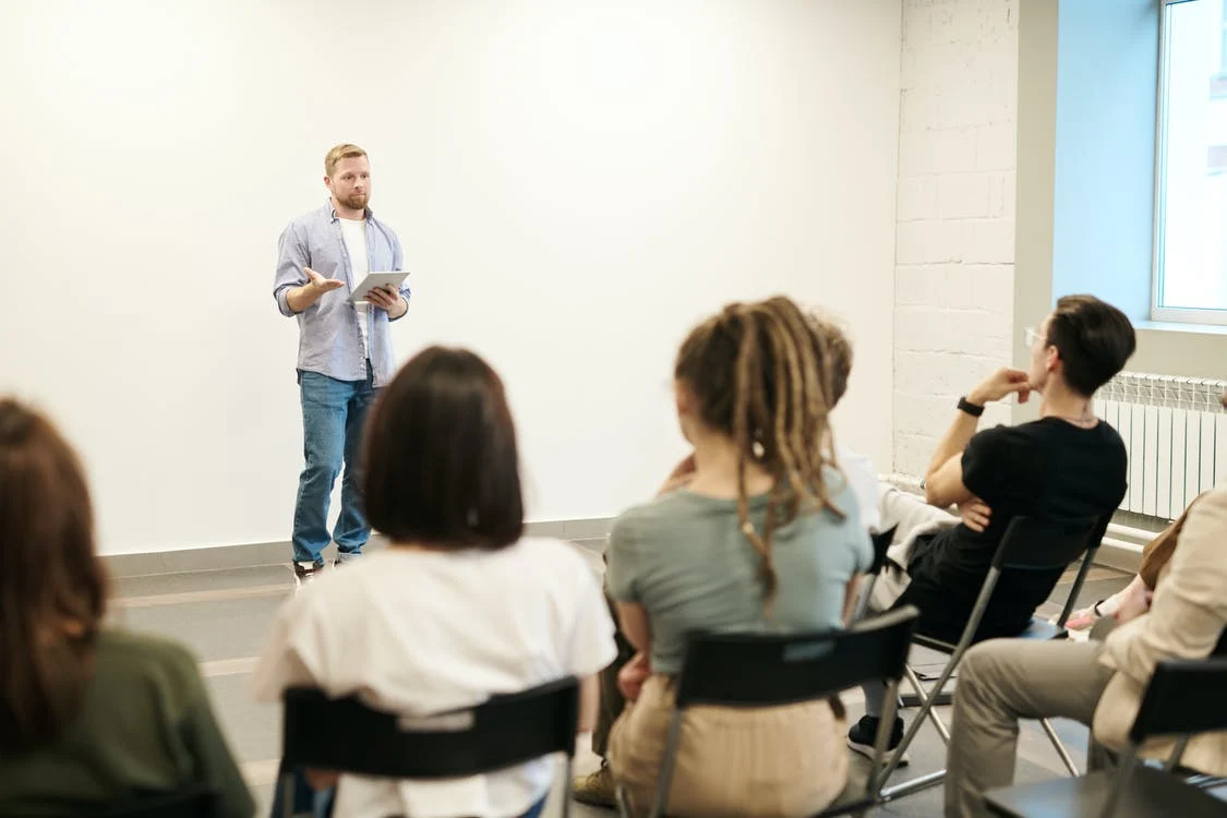 La imagen muestra a un hombre frente a una clase en la que está haciendo una presentación oral a sus alumnos que están sentados y prestando atención. 