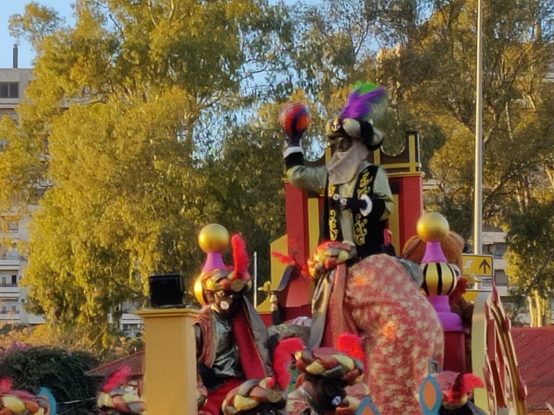 La imagen muestra una cabalgata de rey mago. Montado en ella aparece el rey Baltasar de color, vestido con túnicas marrones y negras y con un sombrero de estilo árabe con plumas moradas y verdes donde se dispone a lanzar al público una pelota roja. 