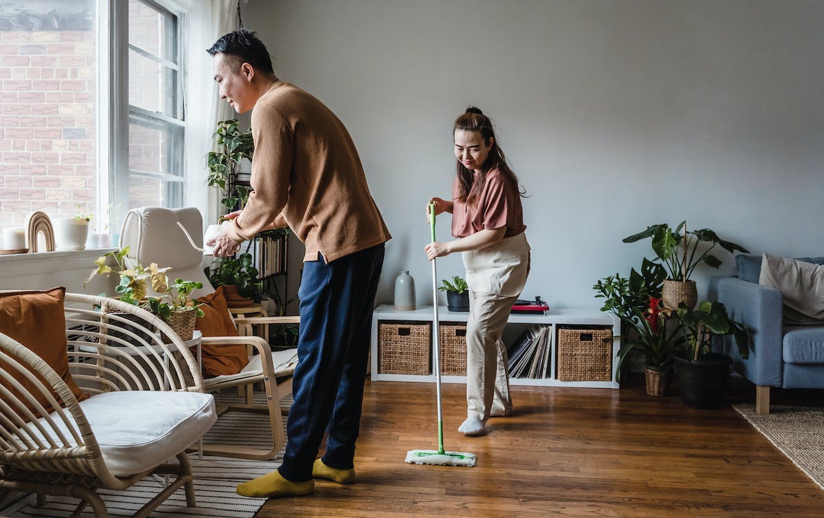La imagen muestra a un hombre y una mujer de origen asiático limpiando la casa. El hombre está a la derecha de la imagen limpiando las hojas de una planta y la mujer en el centro está limpiando el suelo con una mopa. 