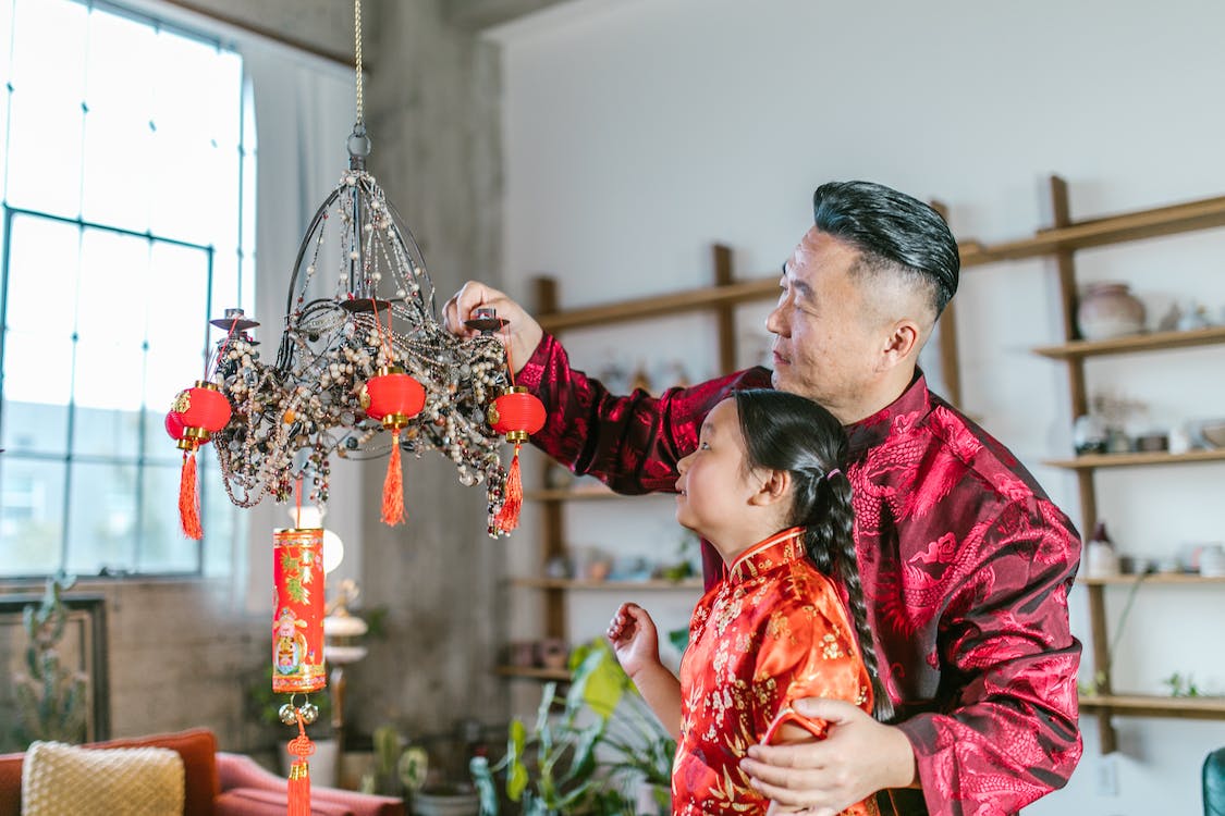 La imagen muestra a un hombre y una niña de origen asiático vestidos con ropa elegante típica china decorando una lámpara con farolillos rojos chinos. 