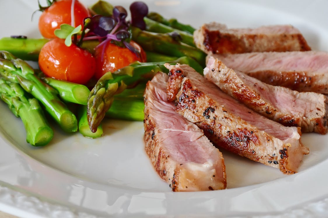 La imagen muestra un plato de comida en la que hay carne de cerdo rosada cortada en láminas acompañado de una ensalada de espárragos verdes y tomates.