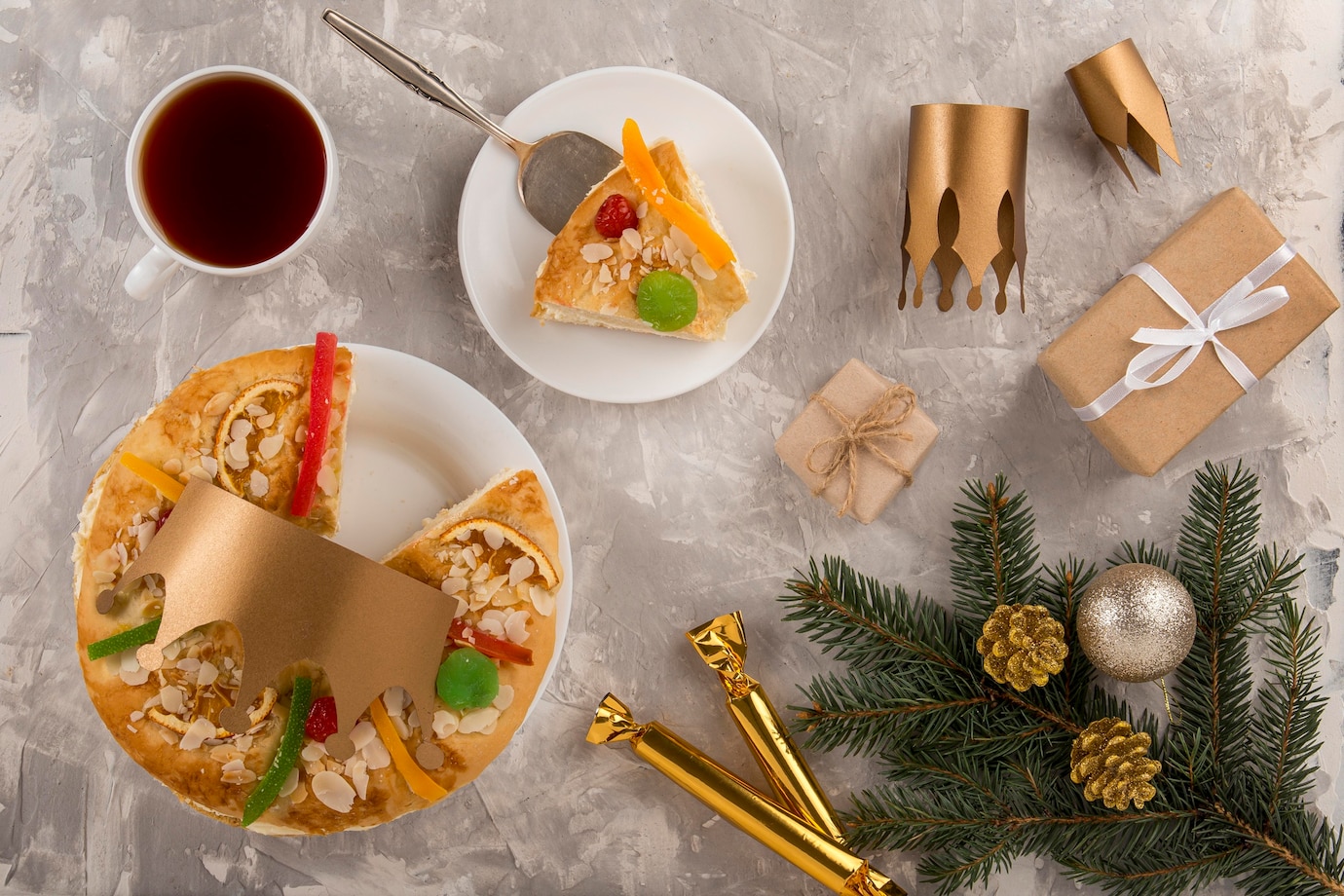 La imagen muestra una mesa con adornos de navidad, una taza de café y un roscón de reyes de color amarillo, en forma de anillo grande y adornado con fruta escarchada y azúcar. 