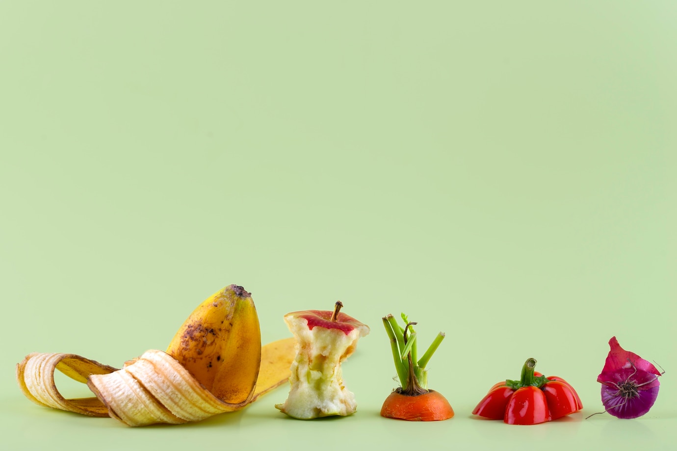 La imagen muestra restos de comida de plátano, manzana, zanahoria, cebolla y pimiento sobre una mesa y con fondo verde agua.