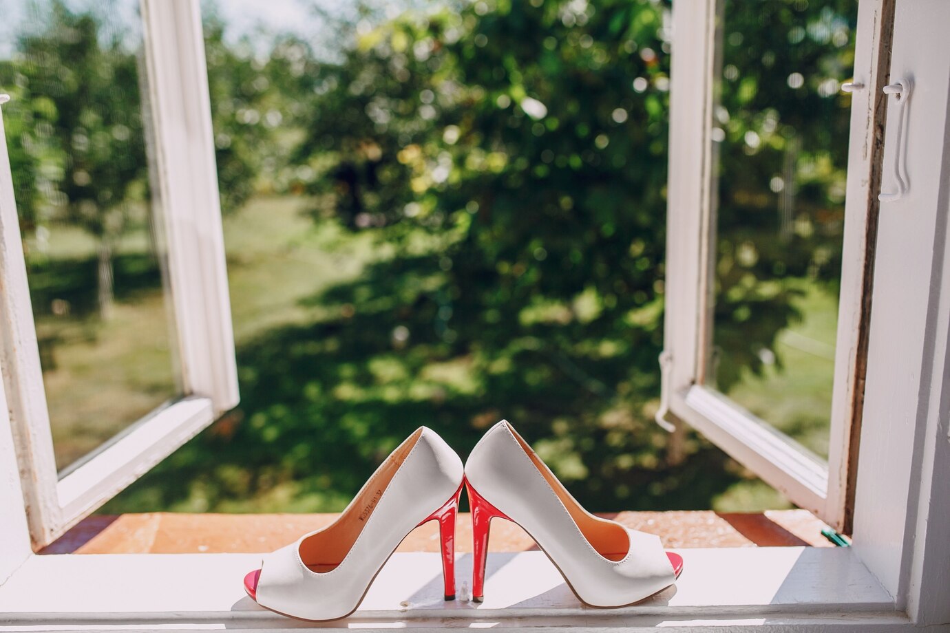 La imagen muestra un par de zapatos de tacón blancos y rojos en la ventana abierta y al fondo se ve un parque. 