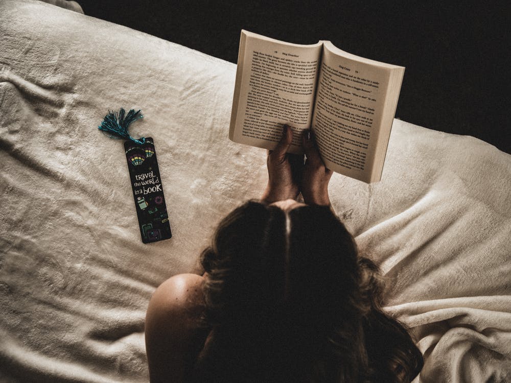 La imagen muestra una mujer tumbada en la cama cogiendo un libro con sus manos y a la izquierda hay un separador de libros.