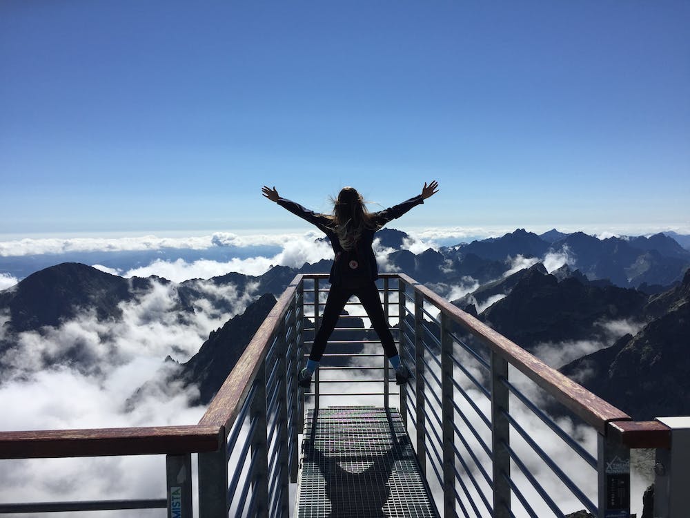 La imagen muestra una chica en un mirador hacia las montañas con los brazos subidos en señal de alegría o victoria. 