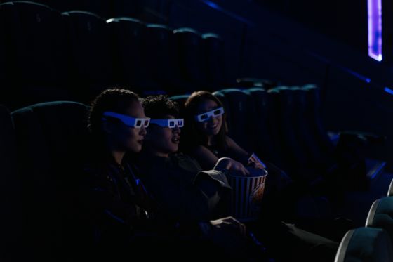 Aparecen tres amigos sentados en el cine disfrutando de una pelicula en con gafas en 3d y comiendo palomitas.