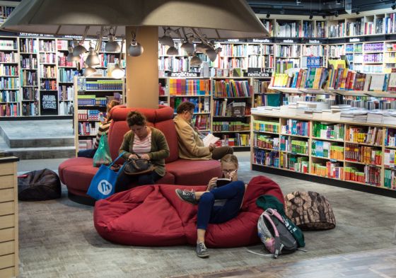 Aparece una biblioteca, en el centro se situa un puff en el que esta leyendo una niña un libro. Detrás hay tres personas leyendo en unos sillones en forma circular.