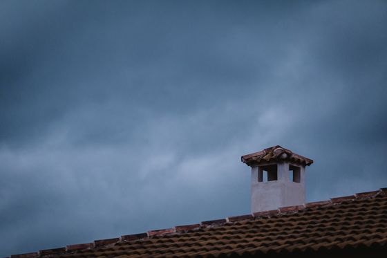 En la fotografía aparece la vista de un tejado de un edificio y  encima del tejado una chimenea de hormigón pintada de blanco con tejas encima de color marrón.