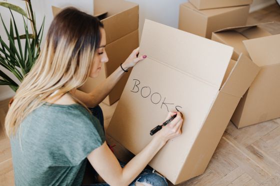 En la fotografía aparece una mujer agachada y de espaldas, sujeta en la mano una caja de carton marron con un nombre puesto. La mujer se situa en posición de leer e identificar lo que hay dentro de la caja.