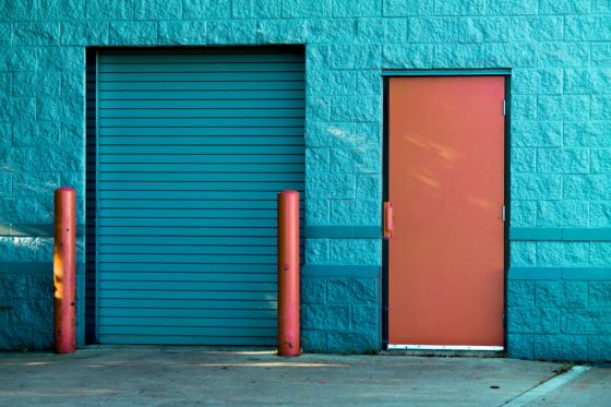 En la fotografía se ve una pared de un edificio pintada de color azul, además tiene una puerta de acceso enrollable pintada en azul y al lado una puerta de hierro pintada de color marrón. .