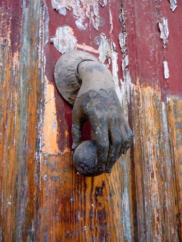La imagen muestra la aldaba en forma de mano de una puerta vieja y deteriorada.